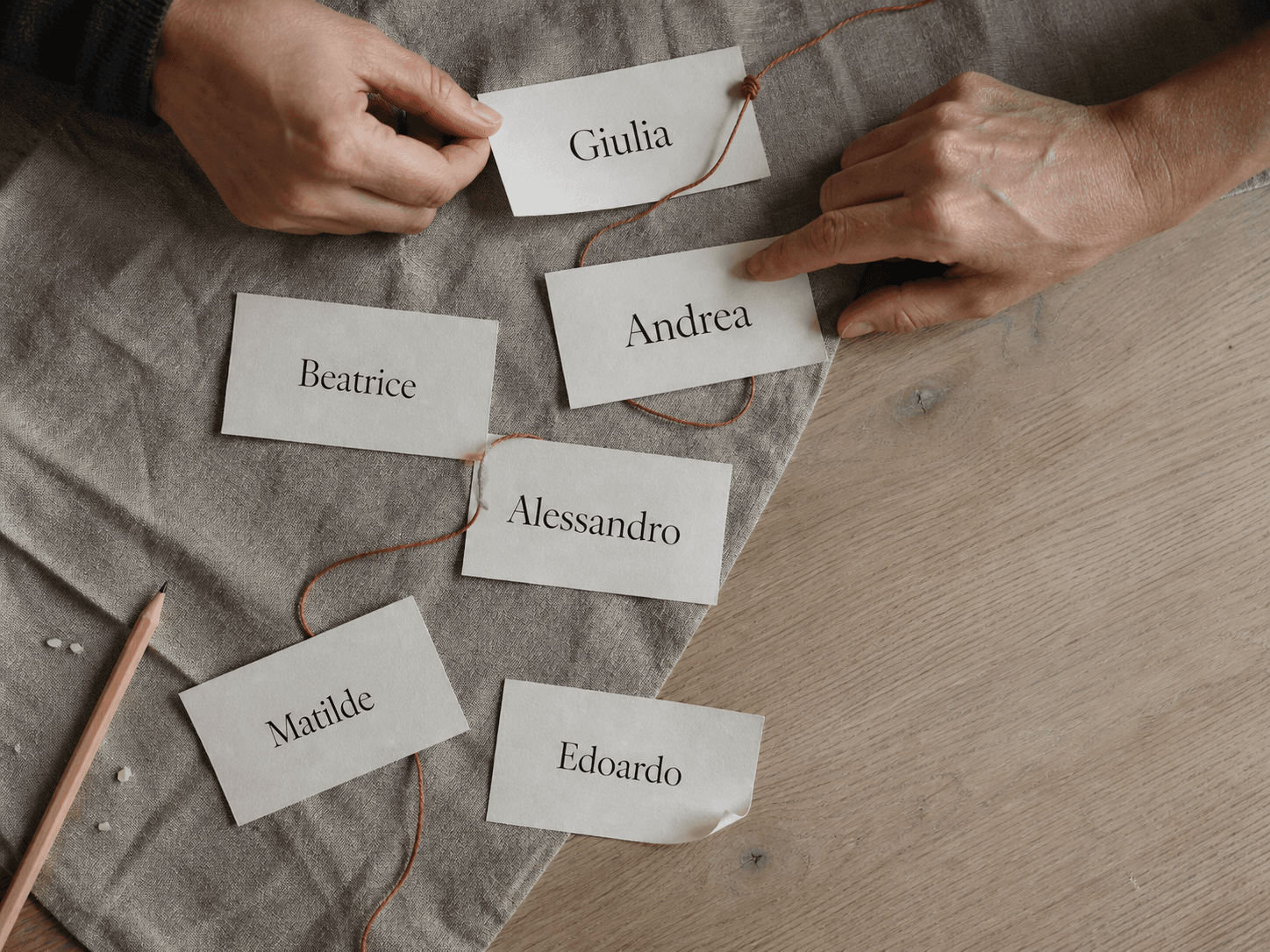 Vista dall'alto: due mani adulte scelgono fra cartellini di carta con i nomi Giulia, Andrea, Beatrice, Alessandro, Matilde ed Edoardo, attraversati da un filo di cotone terracotta